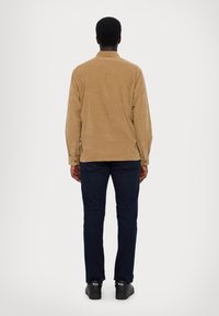 Camicia marrone in velluto a coste con collo e polsini abbottonati, abbinata a jeans blu scuro e scarpe nere con un logo bianco sul tallone.