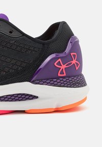 Scarpa sportiva nera con tomaia in rete, supporto per il tallone viola, dettagli rosa e suola bianca. Presenta dettagli testurizzati e una suola colorata.