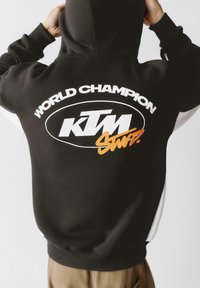 Čierna mikina s kapucňou s bielym textom "SVETOVÝ ŠAMPIÓN" a logom "KTM" v bielej a oranžovej farbe, s voľným strihom a elastickými manžetami.