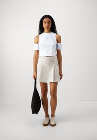 Λευκό κοντό τοπ με ανοιχτούς ώμους και μικρό λογότυπο, σε συνδυασμό με μια ανοιχτόχρωμη πλισέ φούστα και λευκά αθλητικά παπούτσια με καφετί σόλες.