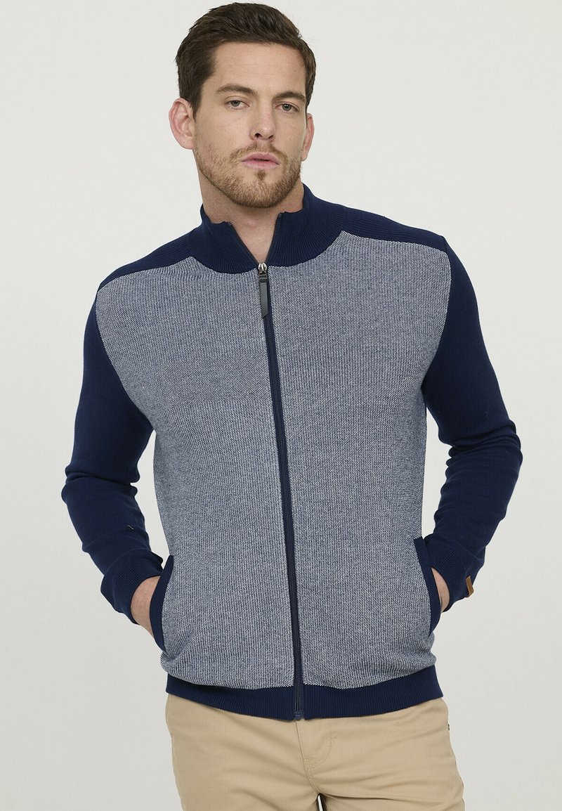 Pull zipé bleu marine et bleu clair avec un tissu texturé, col et poignets côtelés, poches latérales et silhouette ajustée.