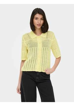 Kvinne iført en kortermet gul heklet topp med knapper og krage, kombinert med høytlivs svarte jeans, mot en ensfarget bakgrunn.