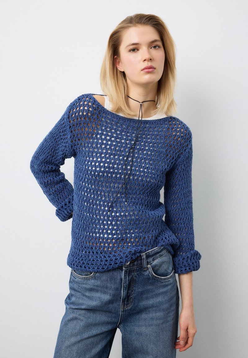 Junge Frau trägt einen blauen, locker gestrickten Pullover über einem weißen Oberteil, blaue Jeans und ein schwarzes Kordhalsband, steht vor einem weißen Hintergrund.