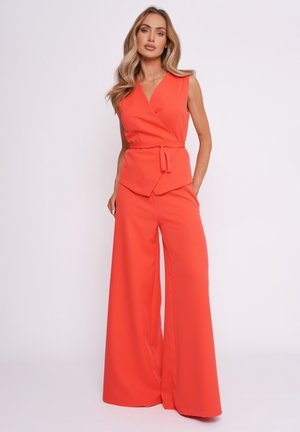 Kobieta ubrana w bezrękawnikowy, jaskrawo pomarańczowy top z kopertowym dekoltem oraz pasujące szerokie spodnie, pozująca na jednolitym tle.