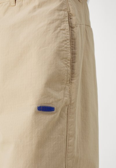 Shorts beiges en fabric léger avec un motif de grille texturé. Présentent une patch logo bleue étiquetée "LES BENJAMINS" sur le côté.
