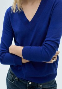 Maglione a V, maniche lunghe in blu reale, realizzato in morbido materiale a maglia. Polsini e orlo a costine, indossato sopra jeans in denim scuro. Design minimalista.