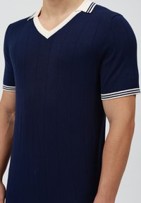 Navy gestricktes Poloshirt mit V-Ausschnitt, geripptem Textil und weißen Streifen am Kragen und den Ärmelabschlüssen. Verfügt über vertikale Streifen.
