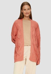 Texturiertes, offen gestricktes Cardigan in Koralle mit lockerer Passform, einer Zopfmusterung und weiten Ärmeln. Kombiniert mit einem beigen Oberteil und hellen Hosen.