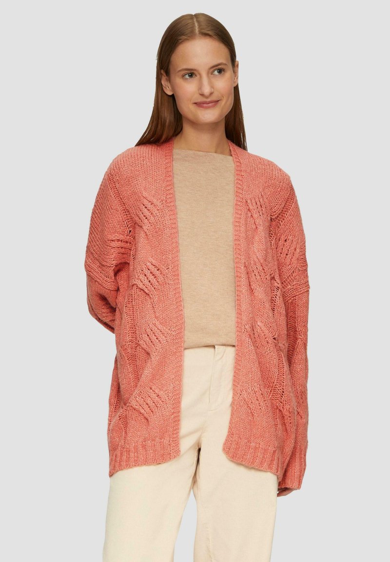 Texturiertes, offen gestricktes Cardigan in Koralle mit lockerer Passform, einer Zopfmusterung und weiten Ärmeln. Kombiniert mit einem beigen Oberteil und hellen Hosen.