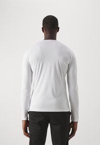 Chemise blanche à manches longues avec une texture lisse, un col rond et une coupe ajustée. L'ourlet est droit et elle présente une légère élasticité.