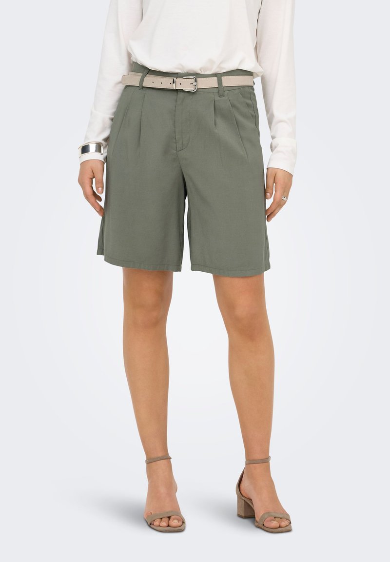 Femme portant un short vert olive ceinturé, un haut blanc à manches longues, un bracelet manchette argenté et des sandales taupe à talon épais avec brides.