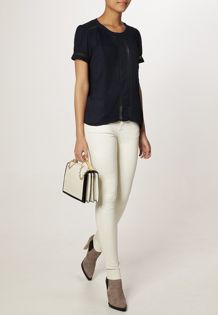 Femme portant un chemisier noir à manches courtes, un pantalon blanc slim, des bottines taupe et tenant un sac à main bicolore avec une anse chaîne dorée.