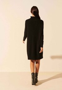 Robe en maille noire avec un col montant, des manches longues et une coupe décontractée, associée à des bottines noires. Texture lisse et longueur au genou.