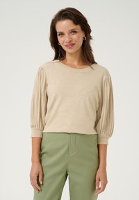 Beige knit pullover with ribbed puff sleeves, round neckline, and lightweight texture. Paired with green high-waisted pants.