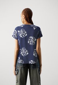 Kvinna med flätat hår som bär stora hoopörhängen, en marinblå kortärmad skjorta med vita blommotiv och urtvättade cargoj jeans, stående vänd bort.