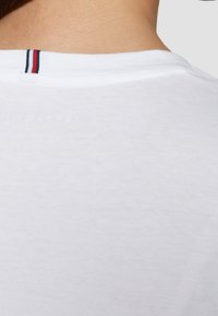 Biała koszulka z okrągłym dekoltem, z kontrastowym pasem w czerwonym i granatowym kolorze z tyłu. Miękka, gładka tkanina o minimalnej teksturze.