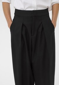 Mustad pleeditud püksid kõrge vöökohaga ja küljetaskutega, valmistatud siledast kangast. Kujundus on puhaste joontega ja kohandatud lõikega.