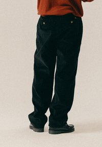 Μαύρο παντελόνι καμπάνα από κορδούρα με χαλαρή εφαρμογή, με χρυσές λεπτομέρειες και πλάγιες τσέπες, συνδυασμένο με μαύρες μπότες.