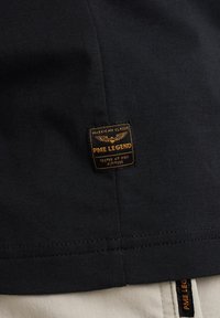Crna pamučna majica s utisnutom PME Legend etiketom narančaste boje. Majica ima glatku teksturu i jednostavan dizajn.