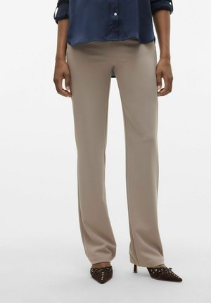 Femme portant un pantalon beige droit, une chemise bleu marine boutonnée et des talons marron foncé à bout pointu avec des clous et un détail nœud.