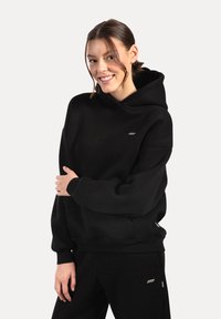 Sweatshirt à capuche noire en tissu doux. Présente des poignets côtelés, une poche kangourou et un petit logo sur la poitrine.