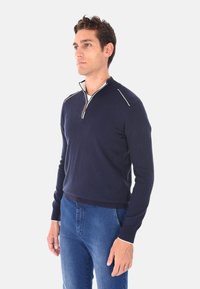 Maglione di lana blu navy con colletto a zip a quarto, dettagli bianchi sulle spalle e sui polsini, caratterizzato da un design aderente e orlo a costine.