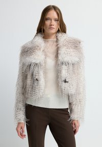 Mujer vestida con una chaqueta de piel texturizada blanca sobre una parte superior de canalé semitranslúcida y pantalones marrón oscuros, de pie contra un fondo blanco liso.