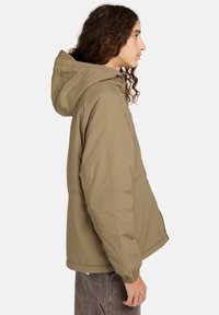 Persoon met lang krullend haar, gekleed in een lichtbruine hoodie en donkere broek, die naar rechts kijkt tegen een effen lichte achtergrond.