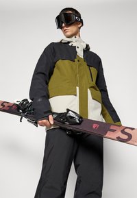 Ski bunda v olivovo zelenej a čiernej farbe, s bielou panelou; obsahuje zipsy, vrecká a kapucňu. Nosič nesie snowboard s viazaním.