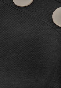 Zwarte ribstof met twee grote, ronde beige knopen op de schouder, met een textuurontwerp en gladde accenten.