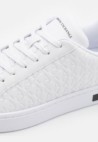 White sneakers with a textured fabric upper featuring a diamond pattern, flat laces, and a black accent on the sole. Logo on the tongue.