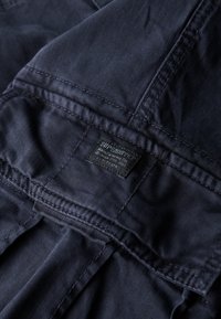 Marineblauwe stoffen label met geperst tekst. Heeft platte naden en een gestikte zoom, met een logo en verzorgingsinformatie. Gladde textuur.