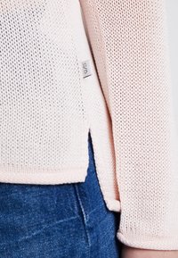 Pull en tricot rose clair avec un tissage lâche, doté d'une étiquette sur le côté et d'un ourlet arrondi, associé à un denim bleu foncé.