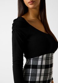 Schwarzes, geripptes Langarmoberteil mit V-Ausschnitt und gerafftem Schulterdetail, kombiniert mit einem karierten Rock in Schwarz- und Grautönen.