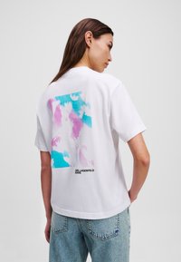 Baltos medvilnės marškinėliai su spalvingu abstrakčiu dizainu mėlyna ir rožine spalvomis nugaroje. Juodas tekstas "Karl Lagerfeld Jeans" apačioje.