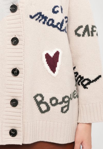 Cardigan lavorato a maglia beige con bottoni neri, motivi testuali in blu navy e verde, e un accento a forma di cuore bianco sul lato sinistro.