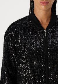 Black bomber jacket with a textured sequin surface. Features a smooth black collar and zipper, showcasing a shiny, eye-catching design.