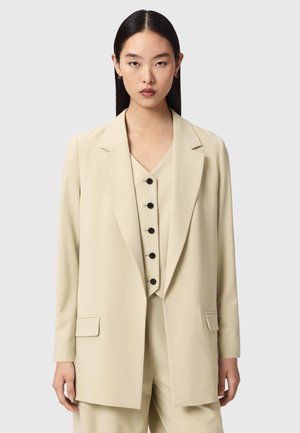 Femme aux cheveux noirs et raides portant un blazer beige ajusté et un gilet assorti boutonné, sur un fond clair uni.