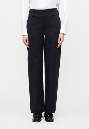 Fekete, szabott, bő szárú nadrágot, fehér hosszú ujjú inget és fekete, fényes mokaszint viselő személy egy egyszerű háttér előtt áll.