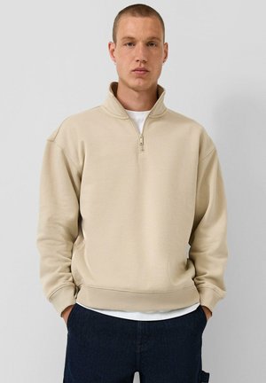 Junger Mann mit kurzen Haaren, der einen beigen Sweatshirt mit Viertelreißverschluss über einem weißen Hemd und dunklen Hosen trägt, steht vor einem schlichten hellen Hintergrund.