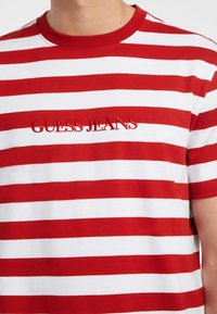 Raudona ir balta dryžuota marškinė su apvalia iškirpte, su iškilmingu logotipu "GUESS JEANS" centrinėje vietoje. Minkšta medvilnė.