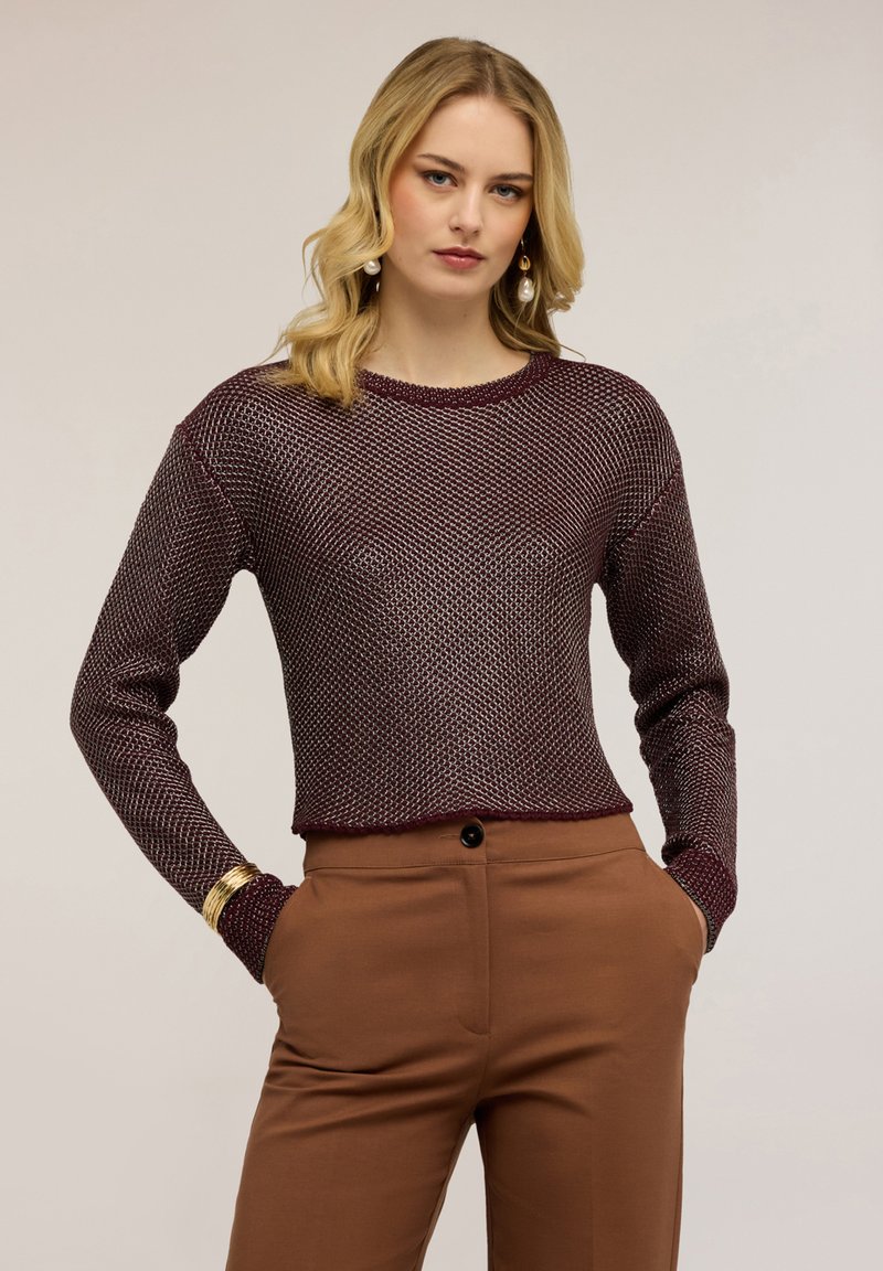 Donna bionda che indossa una maglia a maniche lunghe color granata con trama e pantaloni marroni a vita alta, in piedi con le mani nelle tasche e braccialetti dorati.