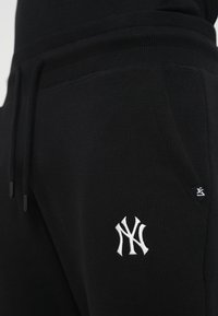 Pantaloni de trening negri cu talie din material ribat, având un logo brodat alb pe coapsă și buzunare laterale. Textură moale, șnur elastic.