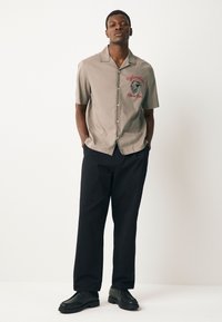 Chemise à manches courtes, gris clair, boutonnée avec un logo sur la poitrine et pantalon noir. La chemise présente un col droit et une texture lisse.