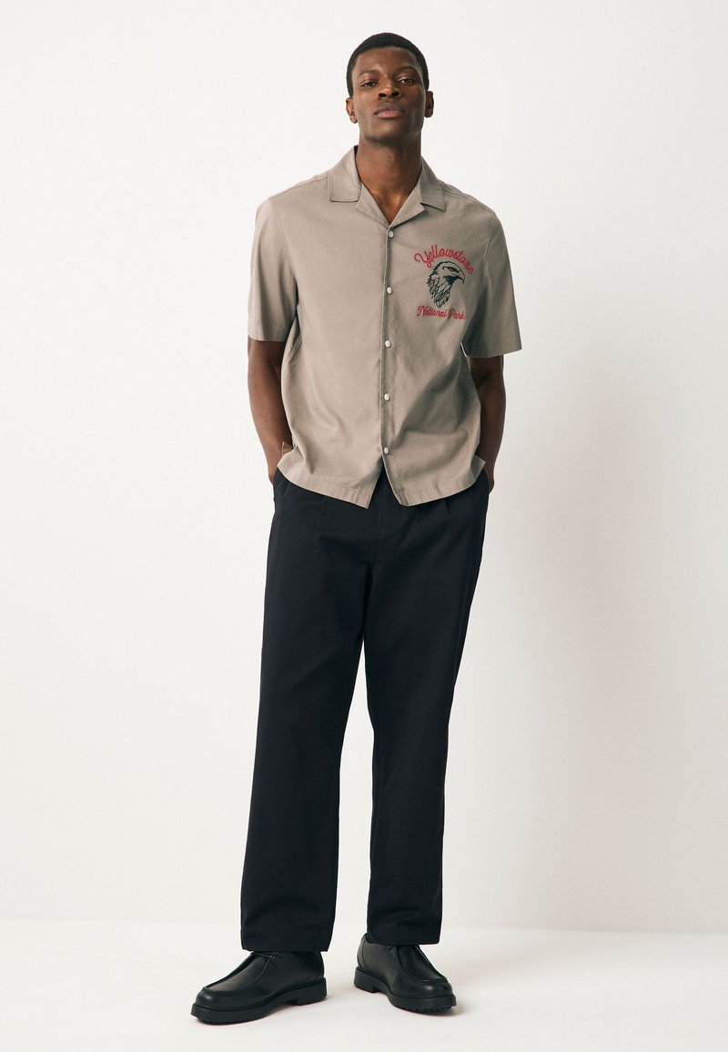 Chemise à manches courtes, gris clair, boutonnée avec un logo sur la poitrine et pantalon noir. La chemise présente un col droit et une texture lisse.