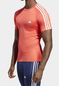 Camisa atlética de manga corta en color coral con tres rayas blancas en los hombros. Hecha de tejido elástico, presenta un diseño ajustado y cuello redondo.