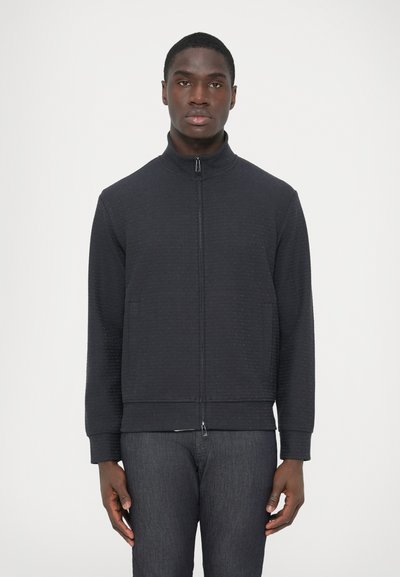 Giacca nera con zip in tessuto strutturato, colletto alto, due tasche frontali e polsini a coste. Indossata con pantaloni in denim scuro.