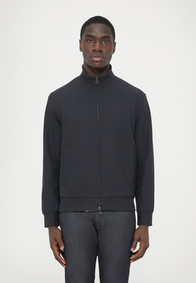 Giacca nera con zip in tessuto strutturato, colletto alto, due tasche frontali e polsini a coste. Indossata con pantaloni in denim scuro.