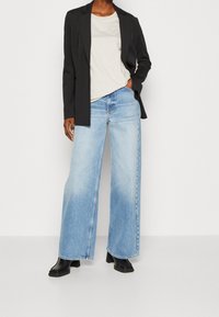 Blazer nero su misura, t-shirt beige con logo, jeans a gamba larga blu chiaro, indossati con stivali neri con tacco. Tessuti morbidi e linee pulite.