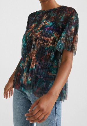 Nærbillede af en person iført en plisseret, flerfarvet bluse med abstrakte mønstre, korte ærmer og lyseblå jeans.